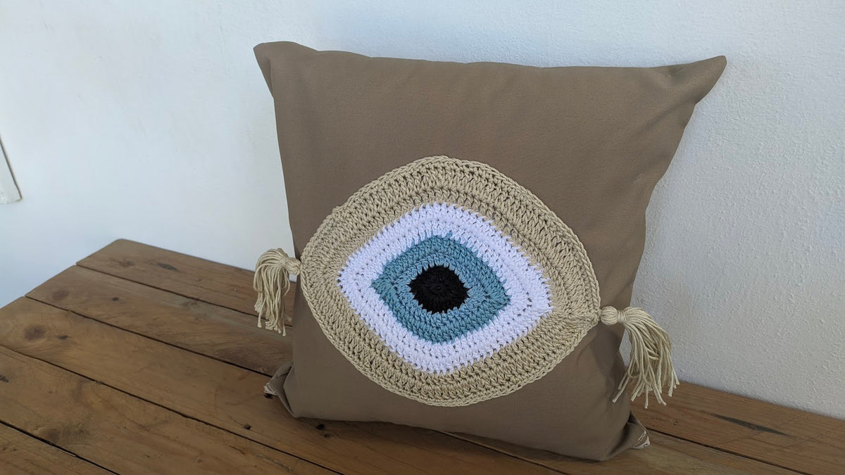 A set of three cushion covers featuring a crochet evil eye design in white, beige, and gray colors with tassels.