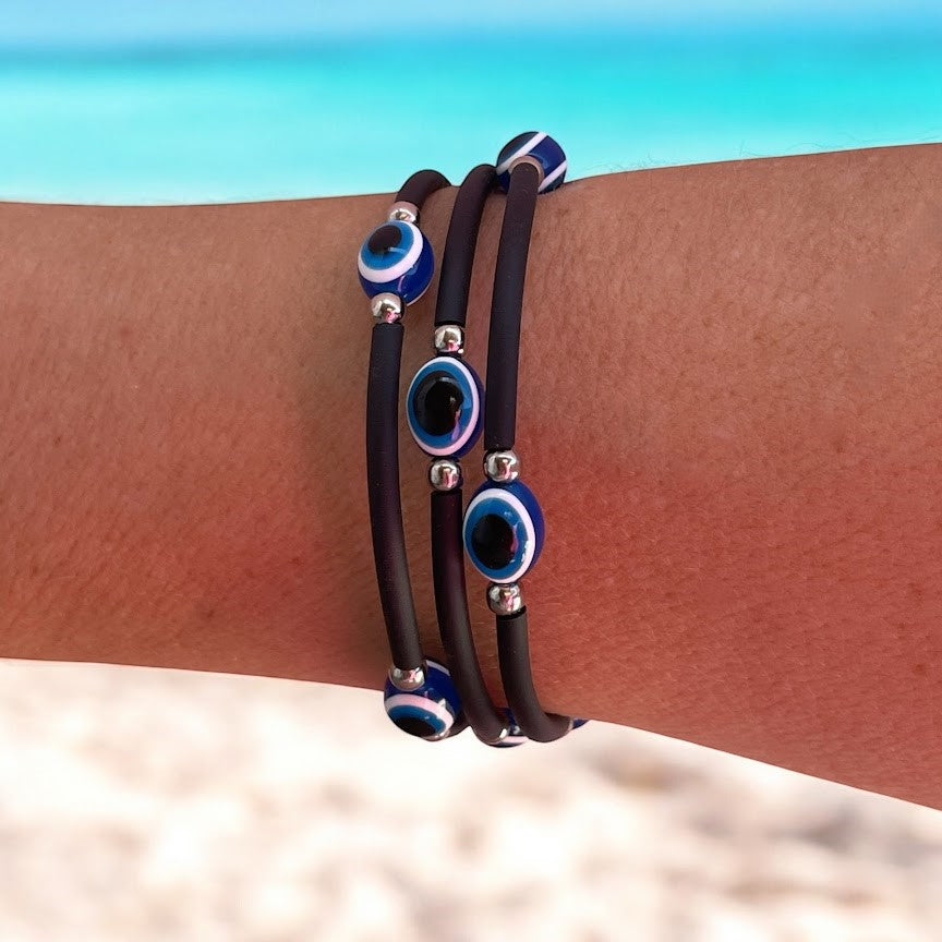 Black rubber bracelets with blue and silver evil eye charms on a wrist against a beach background.