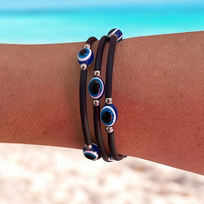 Black rubber bracelets with blue and silver evil eye charms on a wrist against a beach background.