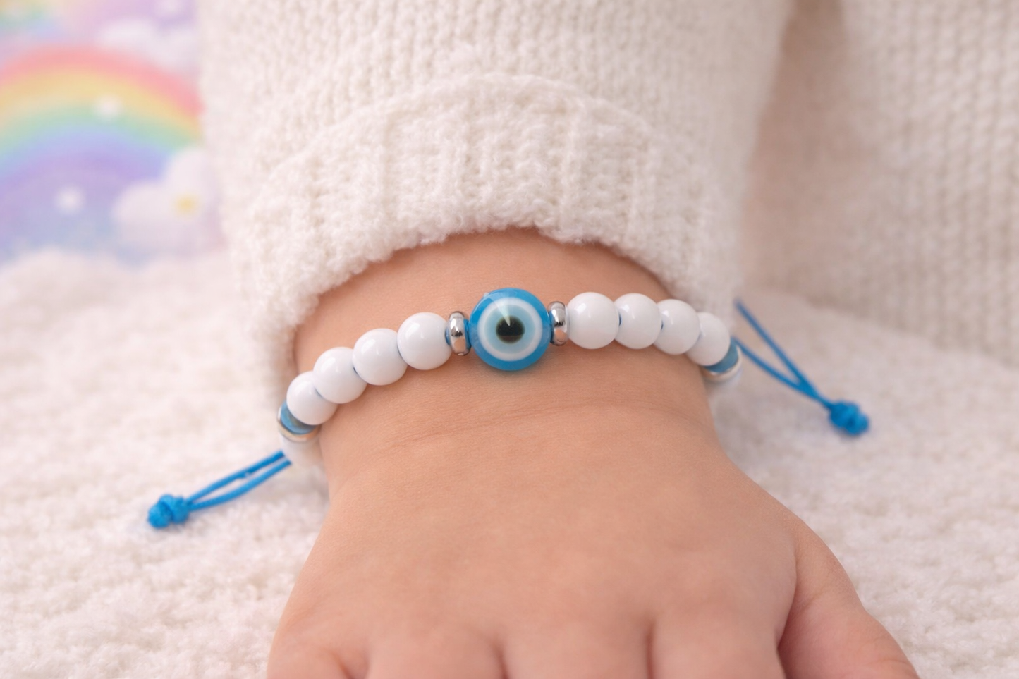 Baby's foot wearing a bracelet with white beads and a blue evil eye charm on a soft surface.