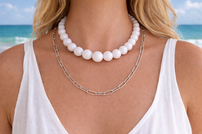 Close-up of a woman wearing a white pearl necklace and silver chain necklace with a beach in the background.
