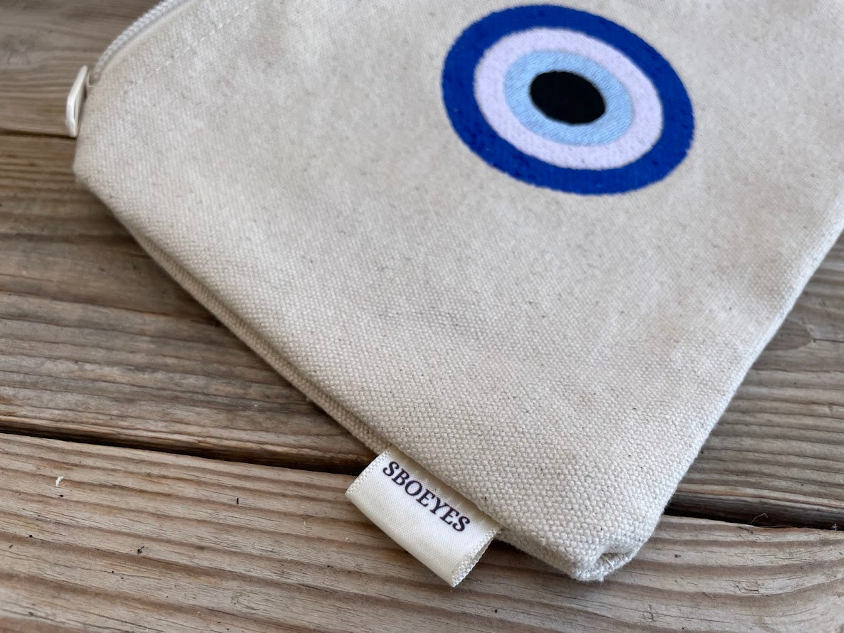 Beige pouch with evil eye design on a wooden surface, featuring the brand 'SBOEYES'.