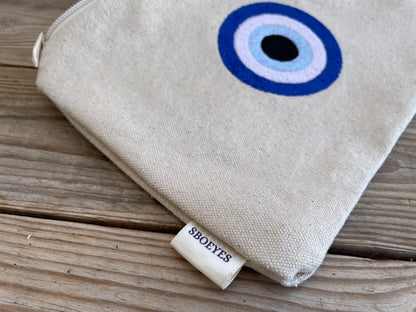 Beige pouch with evil eye design on a wooden surface, featuring the brand 'SBOEYES'.