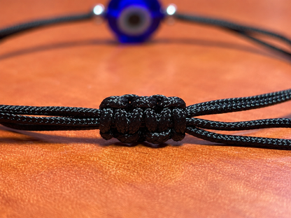 A close-up of a black braided cord bracelet with a knotted central detail, resting on a textured orange-brown surface. A blurred blue evil eye bead is visible in the background.