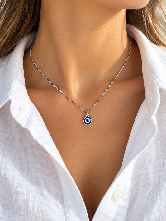 A close-up of a woman wearing a delicate silver chain necklace with a small evil eye pendant. The pendant features concentric circles in blue, white, and black.