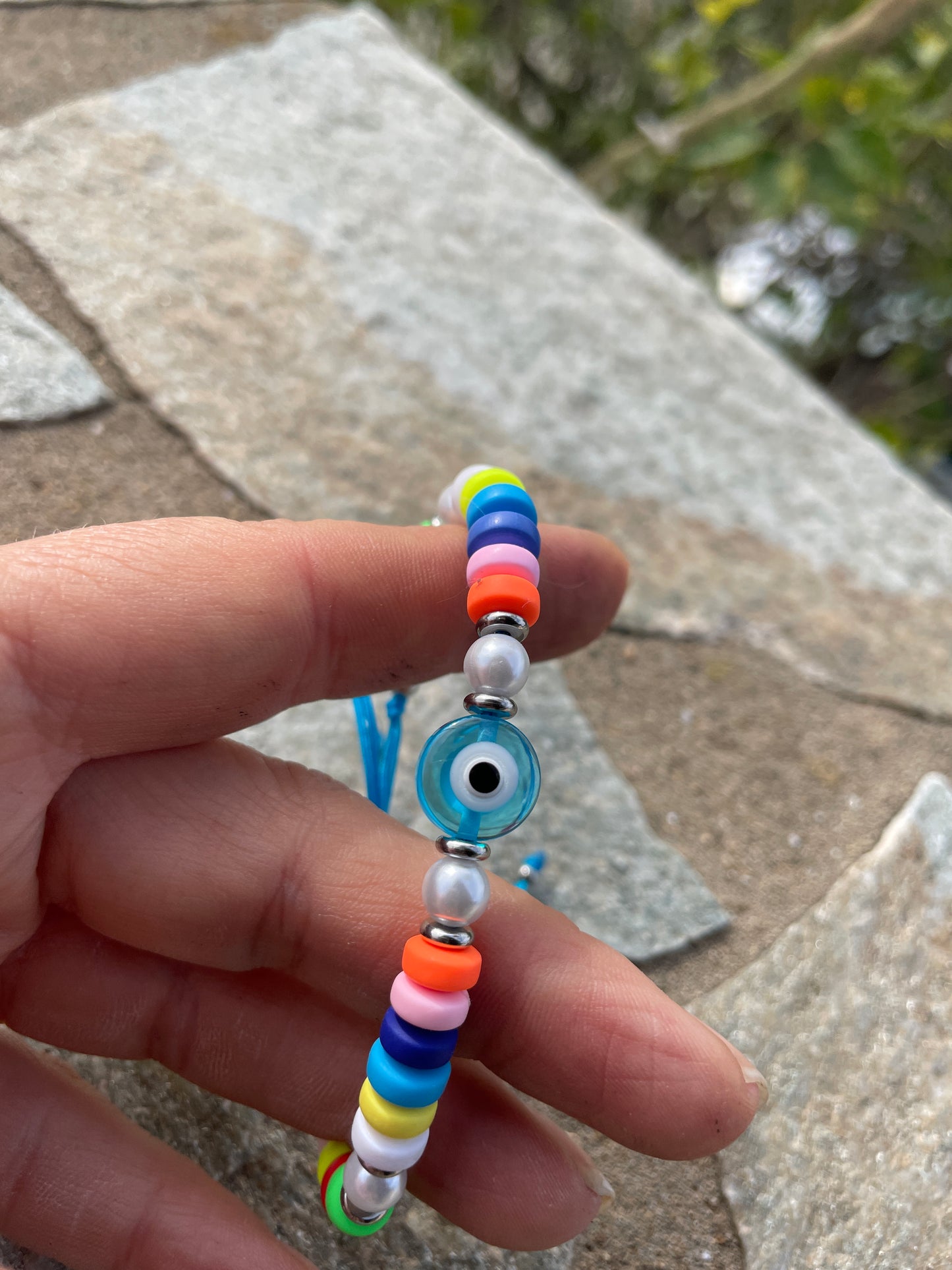 Colorful beaded bracelet with a blue evil eye charm held between fingers on a stone surface.