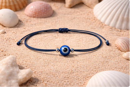 Blue cord bracelet with a silver eye charm on sand with seashells