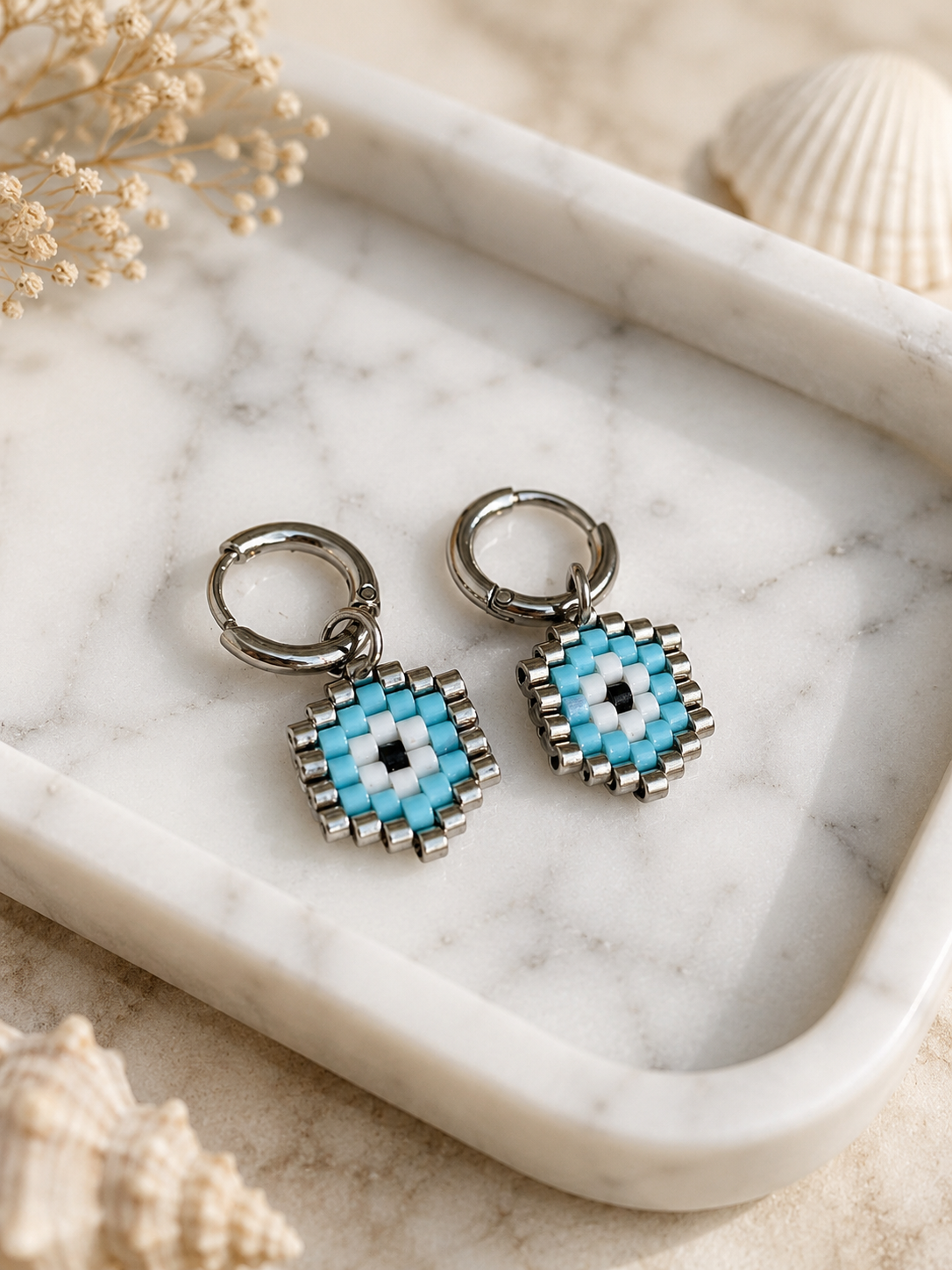 A pair of silver-tone hoop earrings with geometric beaded charms in light blue, white, and black, styled on a white marble tray with dried flowers and seashells.