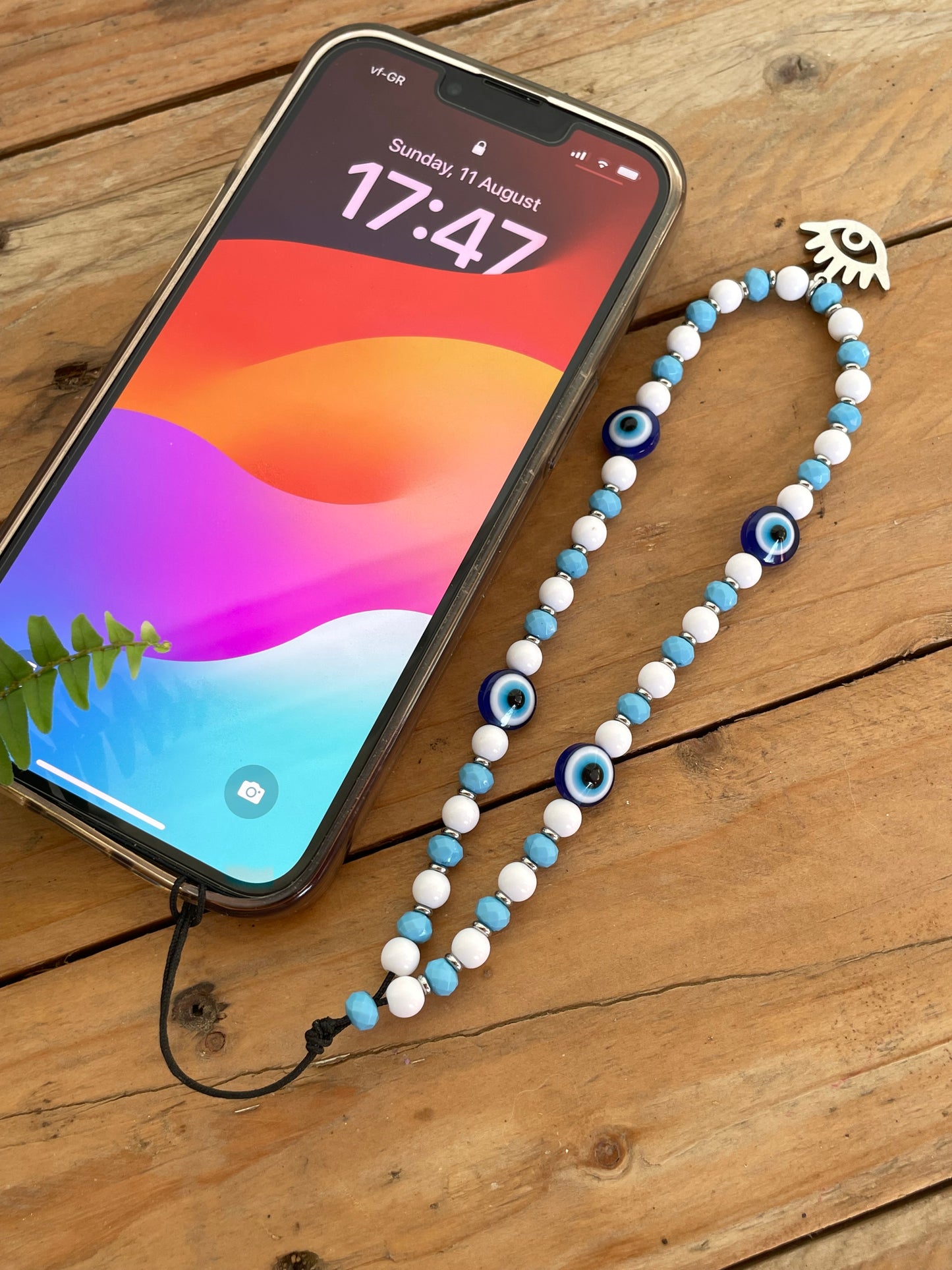 Blue and white evil eye beaded phone strap attached to a smartphone on a wooden table.