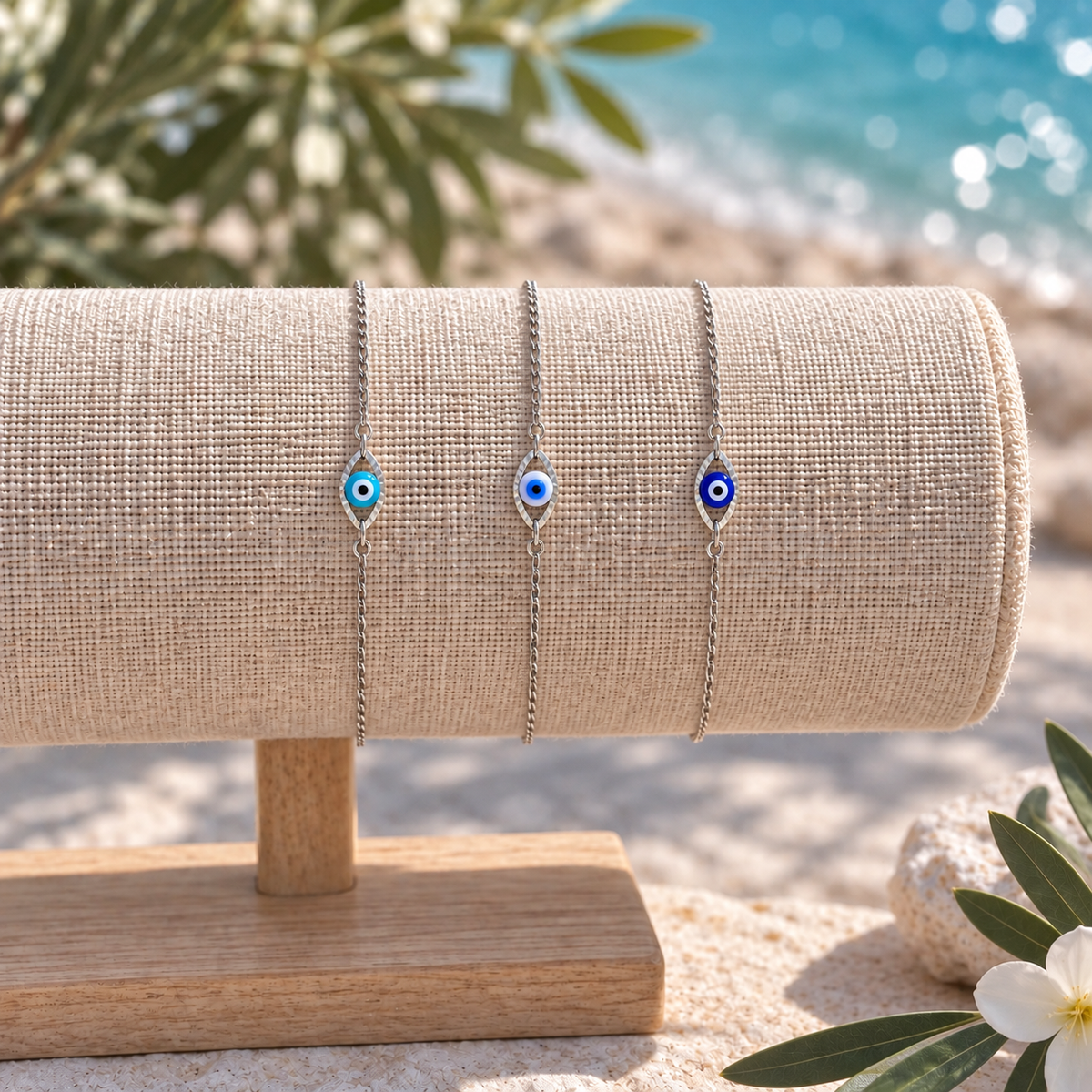 Three delicate silver-tone evil eye bracelets with blue and green accents are displayed on a beige fabric jewelry roll set on a wooden stand, with a blurred beach and olive tree background.