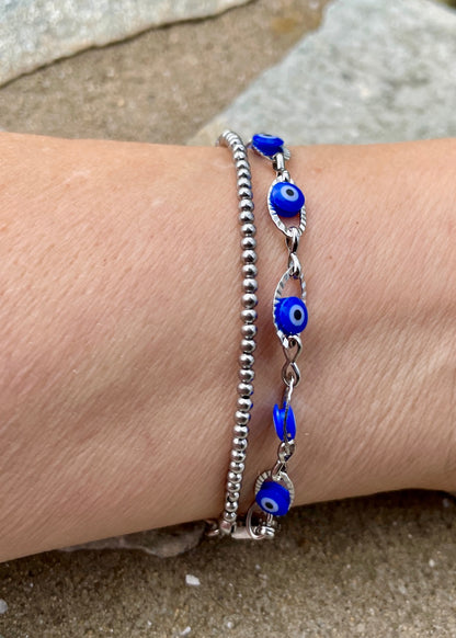Two bracelets with blue evil eye charms on a wrist against a sandy background