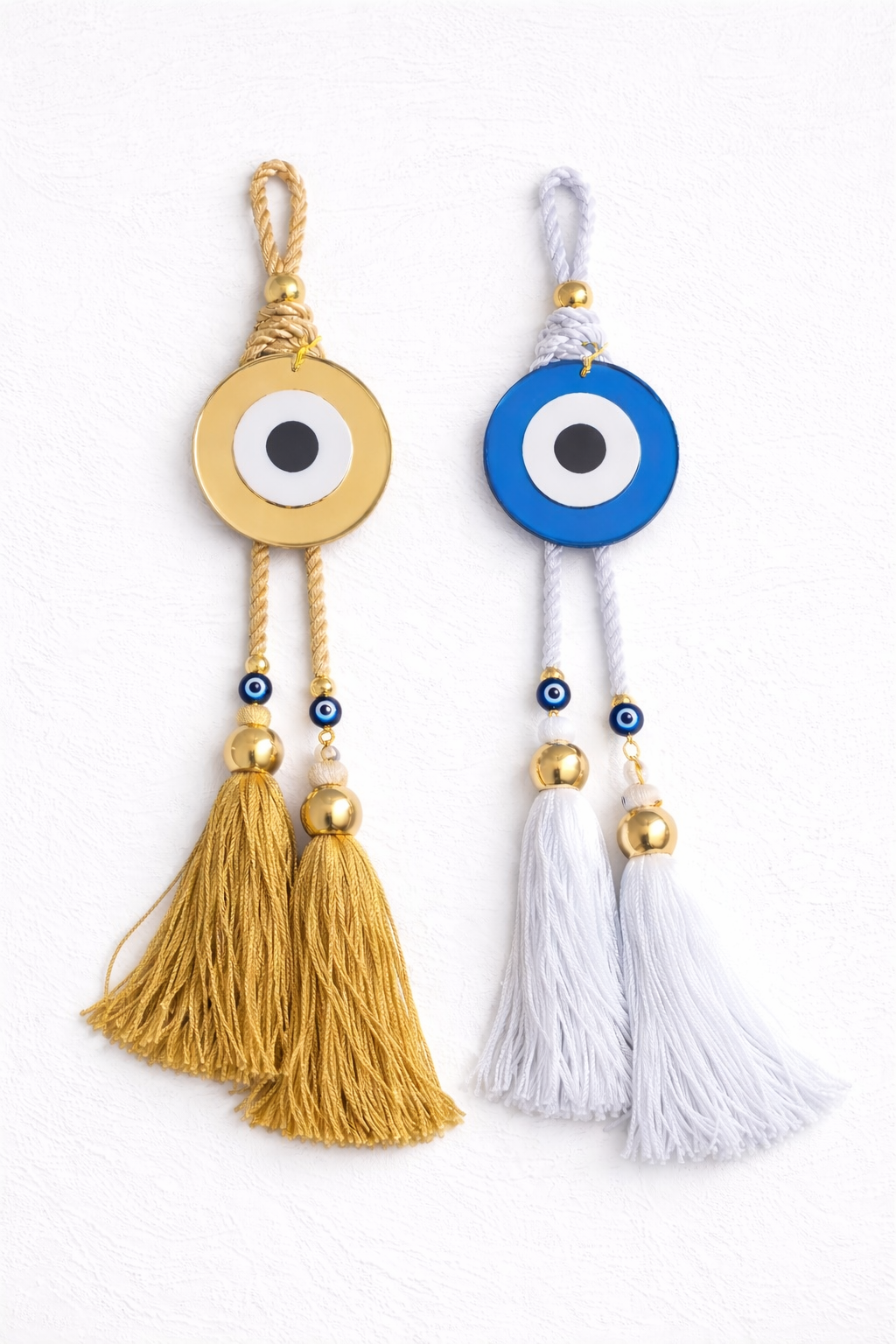 Two evil eye charms with tassels, one gold and one white, on a white background.