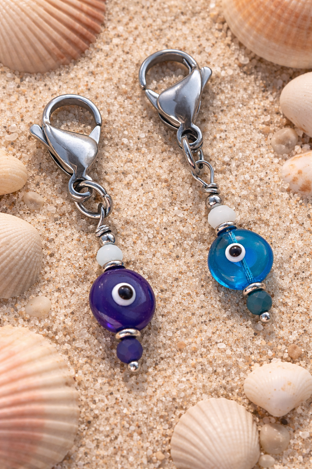 Two silver keychain clips with blue and purple evil eye beads on a sandy background with shells.
