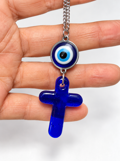 A hand holding a silver chain necklace with a blue glass evil eye charm and a dangling blue glass cross pendant against a white background.