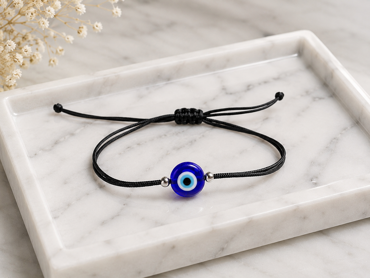 A black adjustable cord bracelet with a blue and white evil eye charm, displayed on a white marble tray.