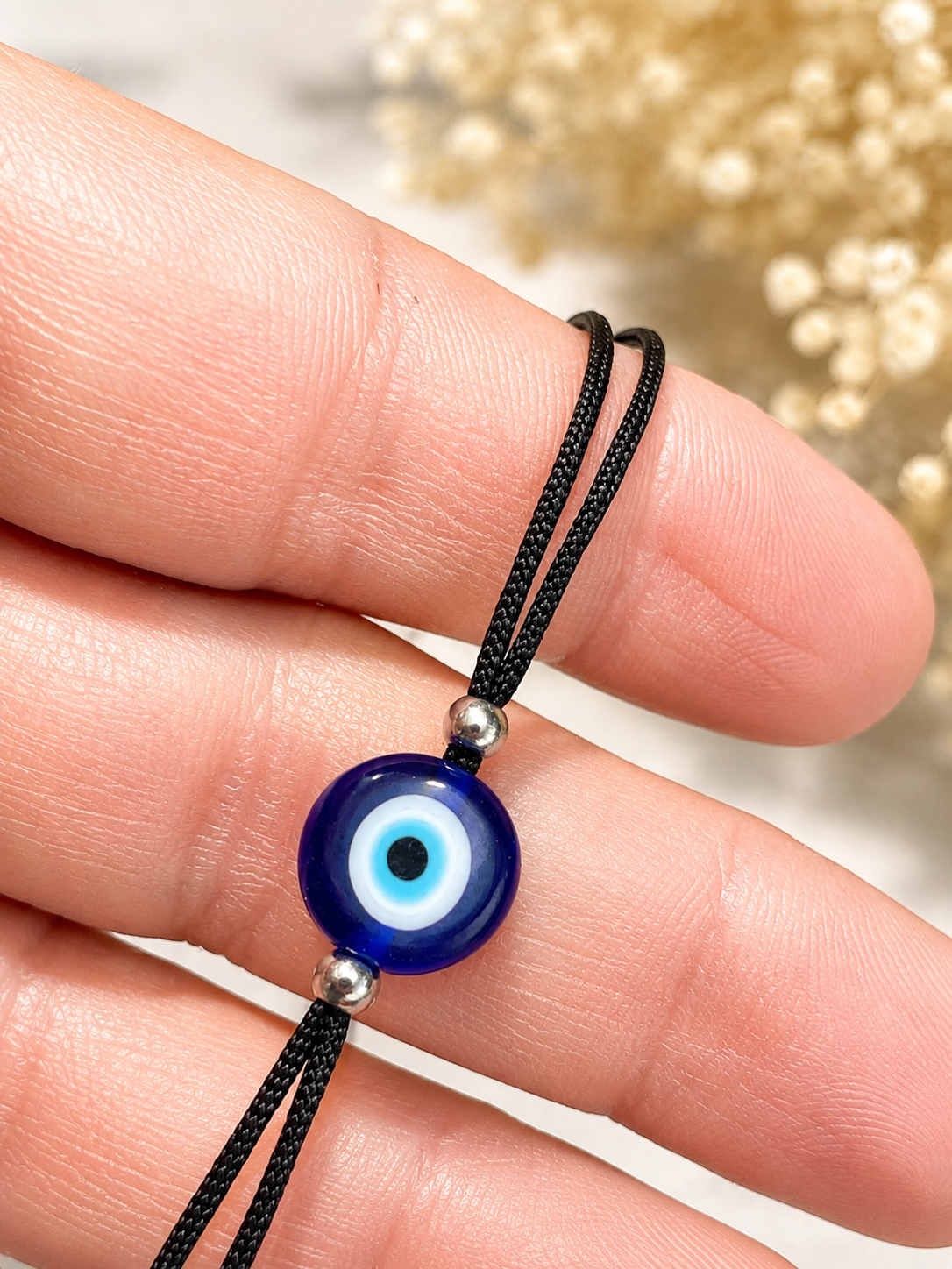 A close-up of a hand holding a black cord necklace with a blue glass evil eye bead and small silver spacer beads, against a softly blurred light background.