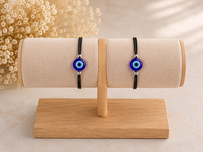 Two black cord bracelets with blue and white evil eye beads displayed on a beige fabric jewelry roll stand on a wooden base.