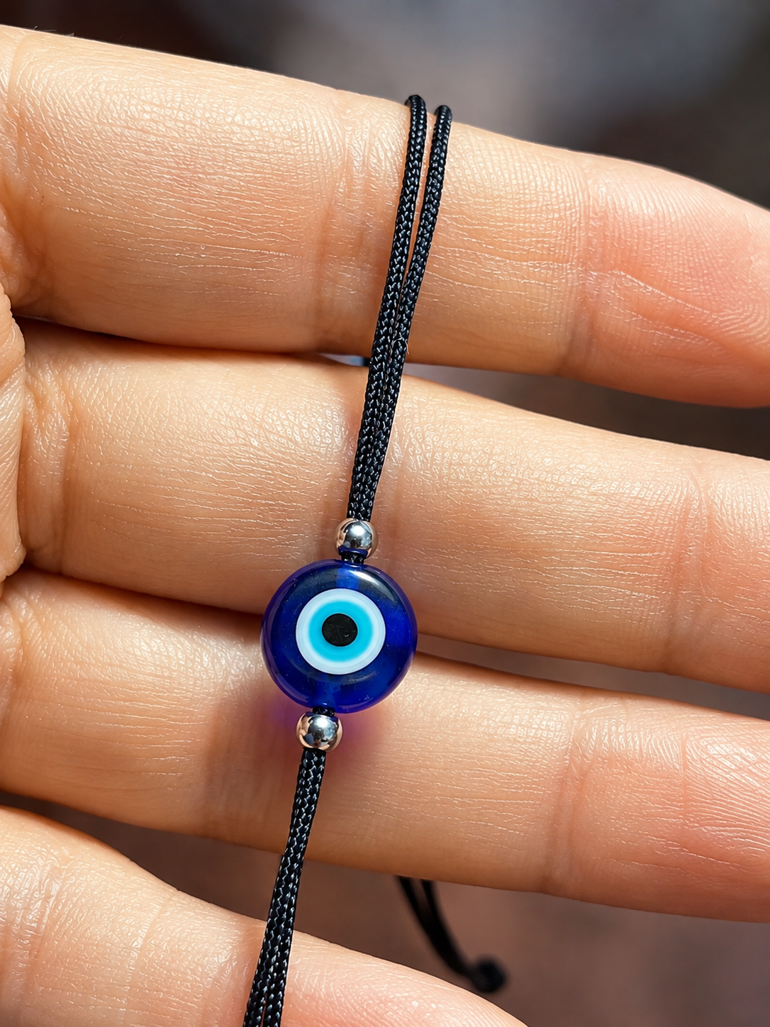 A close-up of a hand holding a black cord bracelet with a blue glass evil eye bead and small silver spacer beads.