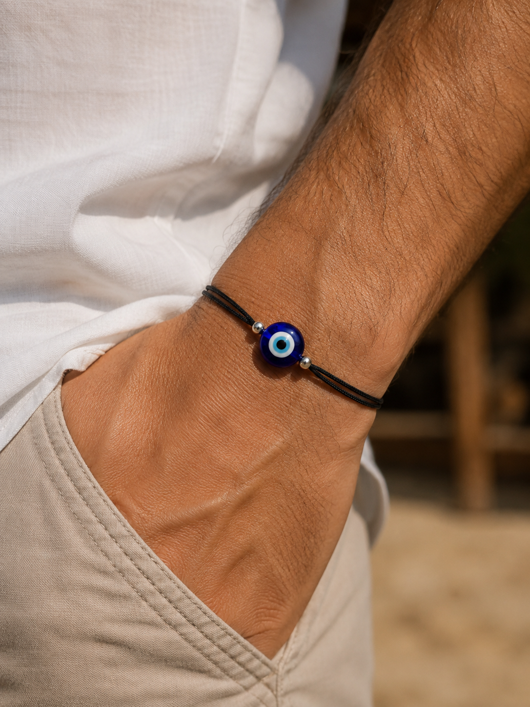 A close-up of a person's wrist wearing a black cord bracelet with a blue and white evil eye charm, paired with a white shirt and beige pants.