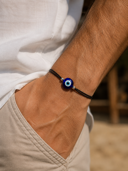 A close-up of a person's wrist wearing a black cord bracelet with a blue and white evil eye charm, paired with a white shirt and beige pants.