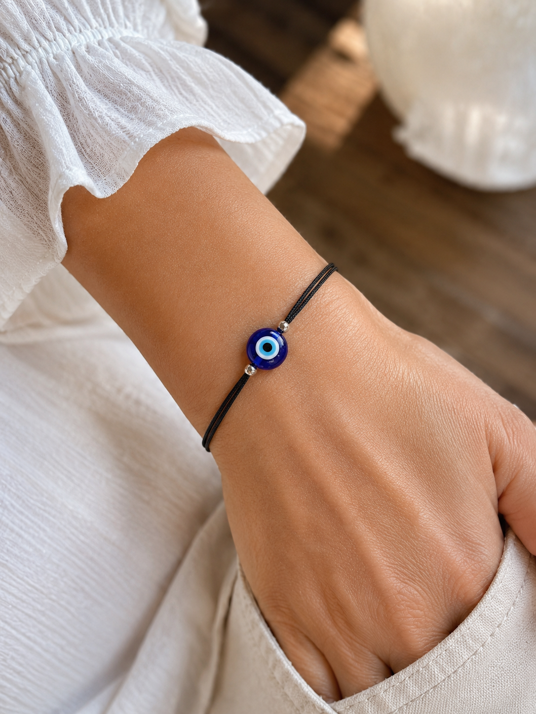 A close-up of a person's wrist wearing a black cord bracelet with a blue and white evil eye charm.