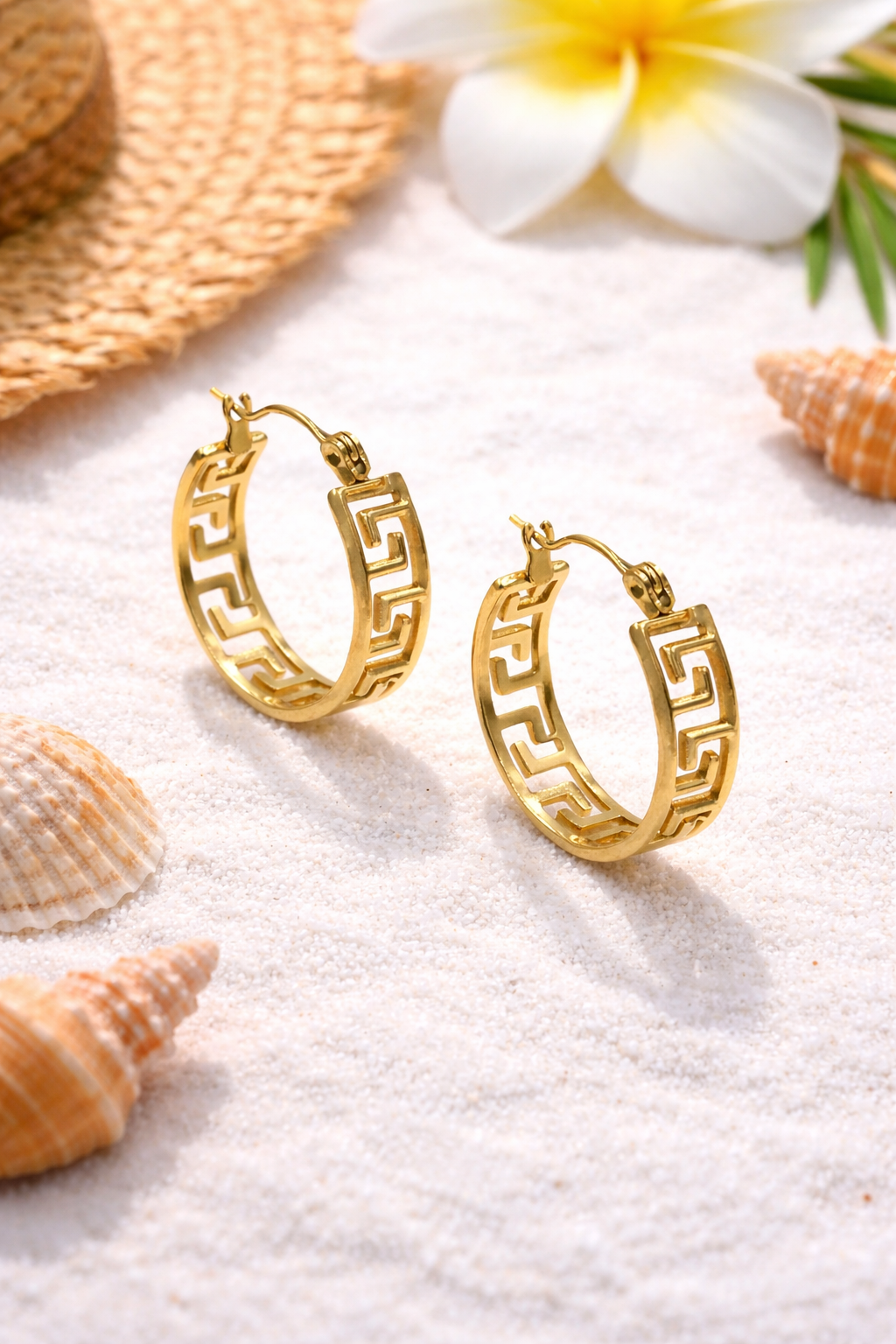 Gold hoop earrings on a sandy surface with shells and flowers