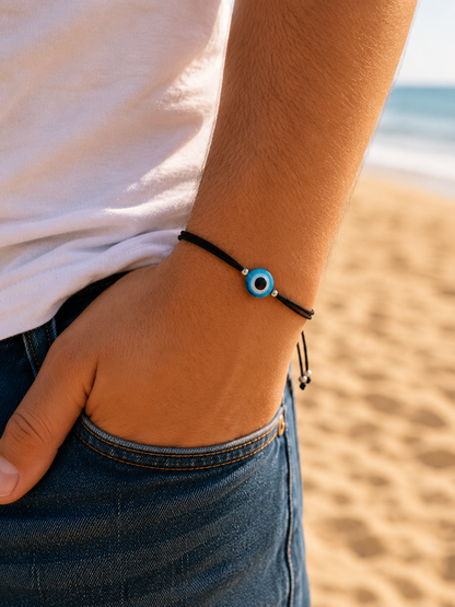 Close-up of an adjustable black cord bracelet with a blue Greek evil eye bead worn on the wrist at the beach.