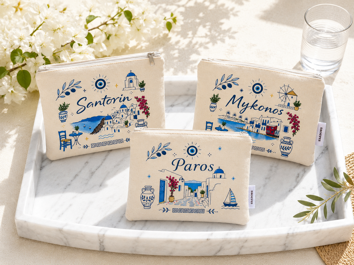 Three pouches with 'Santorin', 'Mykonos', and 'Paros' designs on a marble surface with flowers and a glass of water.