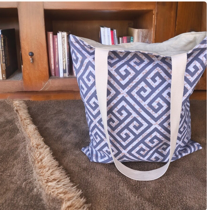 Grey patterned bag with beige straps on a brown carpet next to a wooden cabinet with books.