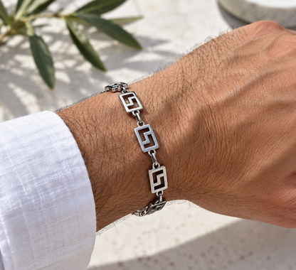 A silver-toned metal bracelet with geometric open-link designs worn on a man's wrist, shown against a light stone background with a white linen shirt cuff visible.