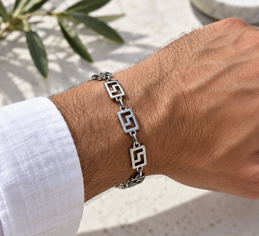 A silver-toned metal bracelet with geometric open-link designs worn on a man's wrist, shown against a light stone background with a white linen shirt cuff visible.