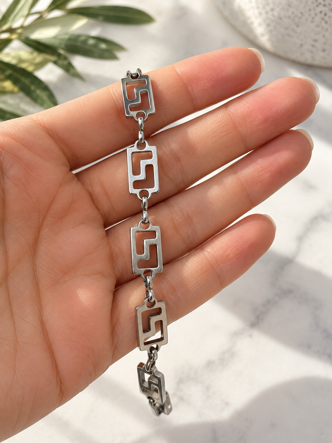 A silver-toned chain bracelet with four rectangular Greek key link charms, held in a person's hand against a light marble background with green leaves in the corner.