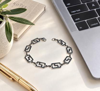 A silver-toned chain bracelet with geometric interlocking links, displayed on a light beige desk surface next to a laptop, a notebook, a pen, and a small potted plant.
