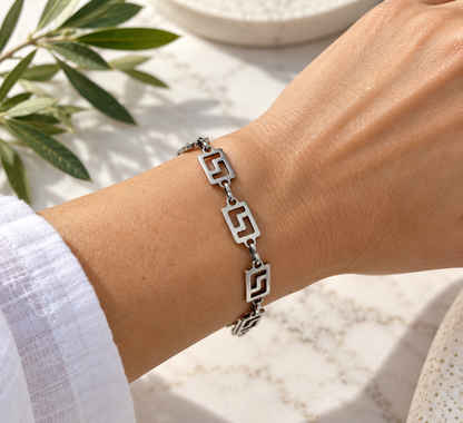 A silver-toned geometric link bracelet with openwork square motifs, shown worn on a person's wrist against a light marble surface with green leaves in the background.