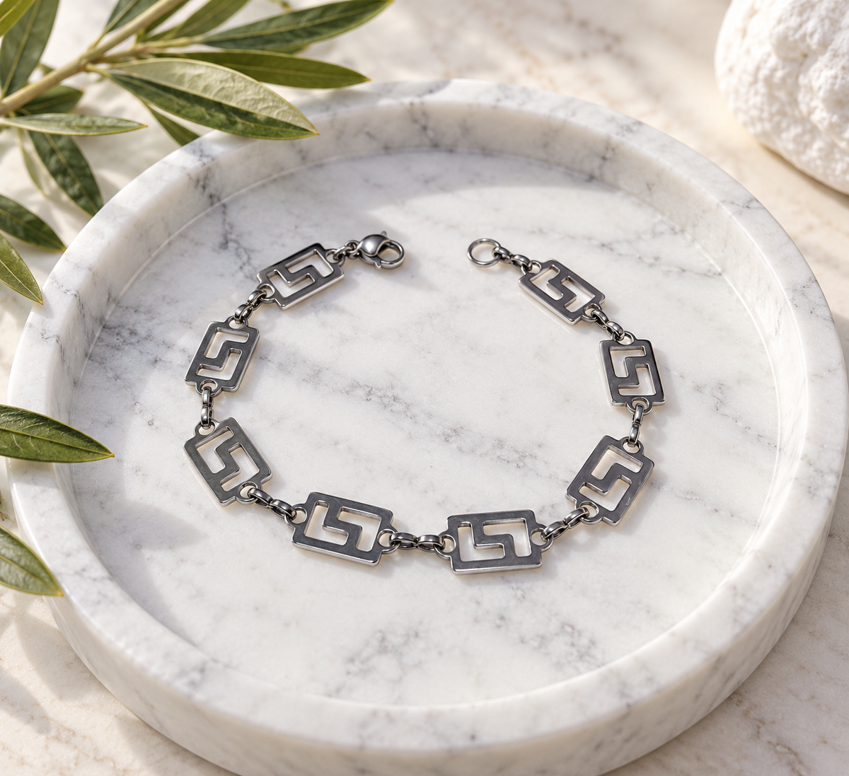 A silver-toned chain bracelet with geometric cutout links, displayed on a round white marble tray with olive leaves and a white stone in the background.