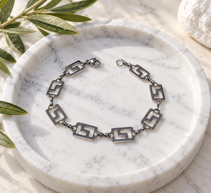 A silver-toned chain bracelet with geometric cutout links, displayed on a round white marble tray with olive leaves and a white stone in the background.
