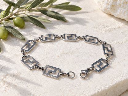 A silver-toned geometric link bracelet with openwork square motifs and a lobster clasp, arranged on a light stone surface with olive branches as styling props.