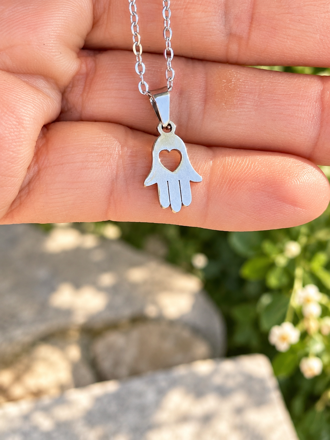 A silver hamsa hand pendant necklace with a heart cutout, held in a person's hand against a blurred outdoor background of green foliage and light-colored stones.
