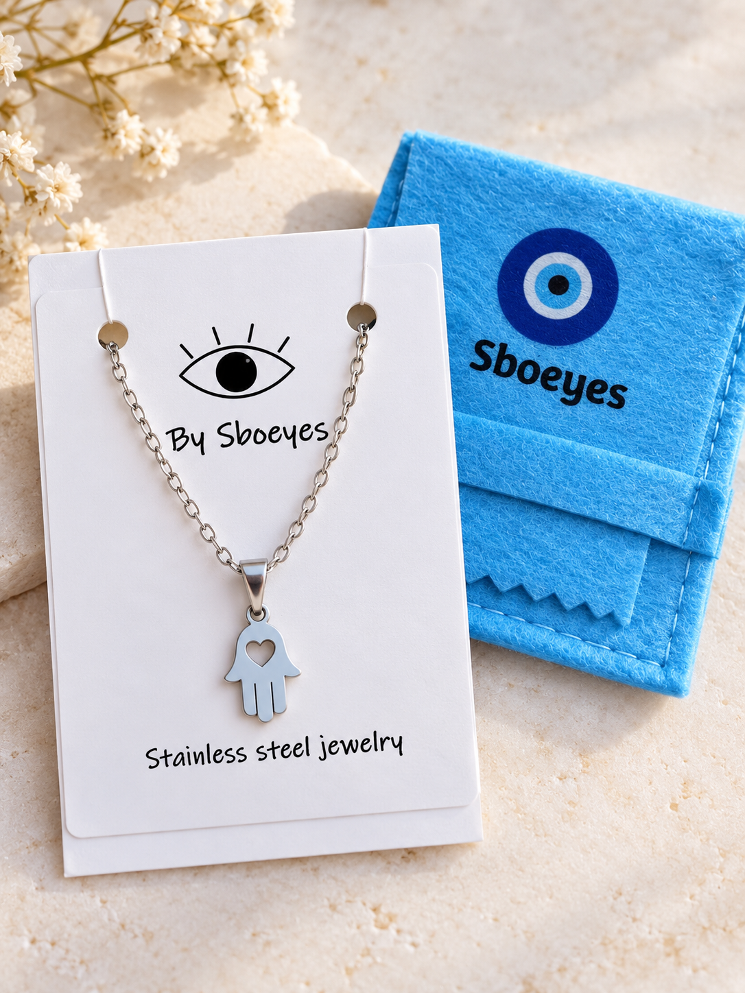 Silver Hamsa hand necklace with a heart detail, displayed on a white card next to a blue felt pouch branded 'Sboeyes' on a light beige textured surface.