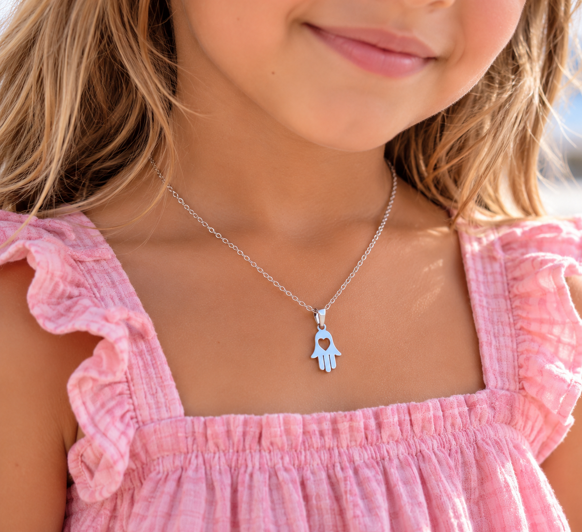 A close-up lifestyle shot of a young girl wearing a silver necklace with a light blue hamsa hand pendant that features a heart-shaped cutout in the center.