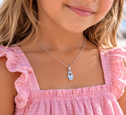 A close-up lifestyle shot of a young girl wearing a silver necklace with a light blue hamsa hand pendant that features a heart-shaped cutout in the center.