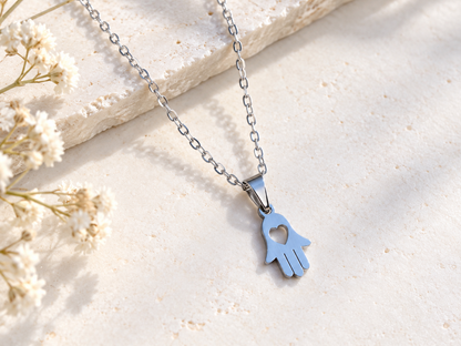 A silver-toned Hamsa hand pendant necklace with a heart cutout, resting on a textured beige stone slab with dried white flowers in the corner.