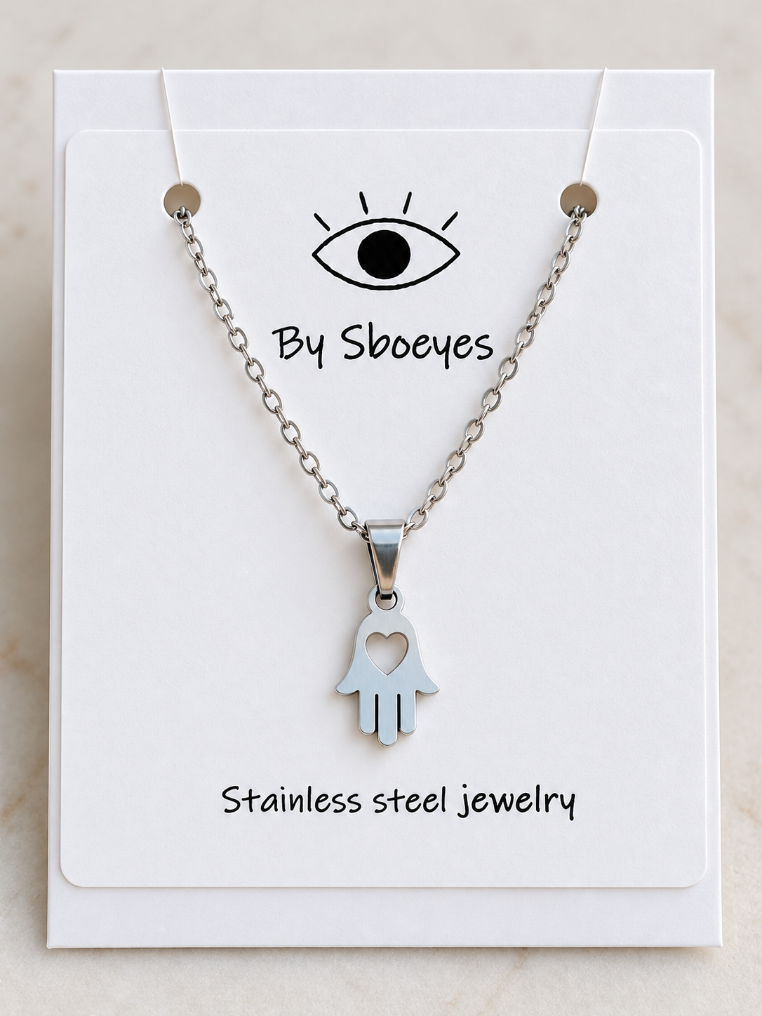 Silver Hamsa hand pendant necklace with a heart cutout, displayed on a white backing card labeled 'By Sboeyes' and 'Stainless steel jewelry'.