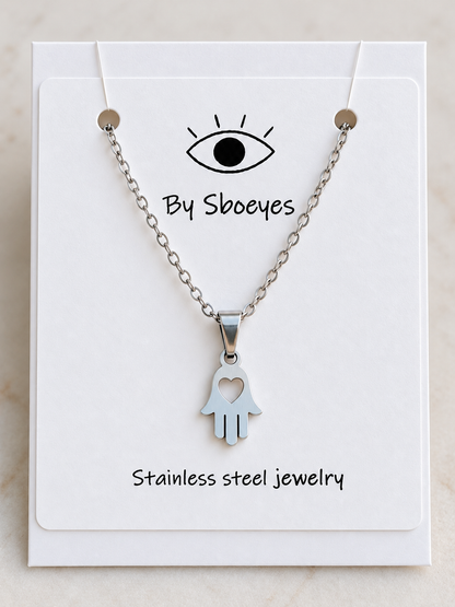 Silver Hamsa hand pendant necklace with a heart cutout, displayed on a white backing card labeled 'By Sboeyes' and 'Stainless steel jewelry'.