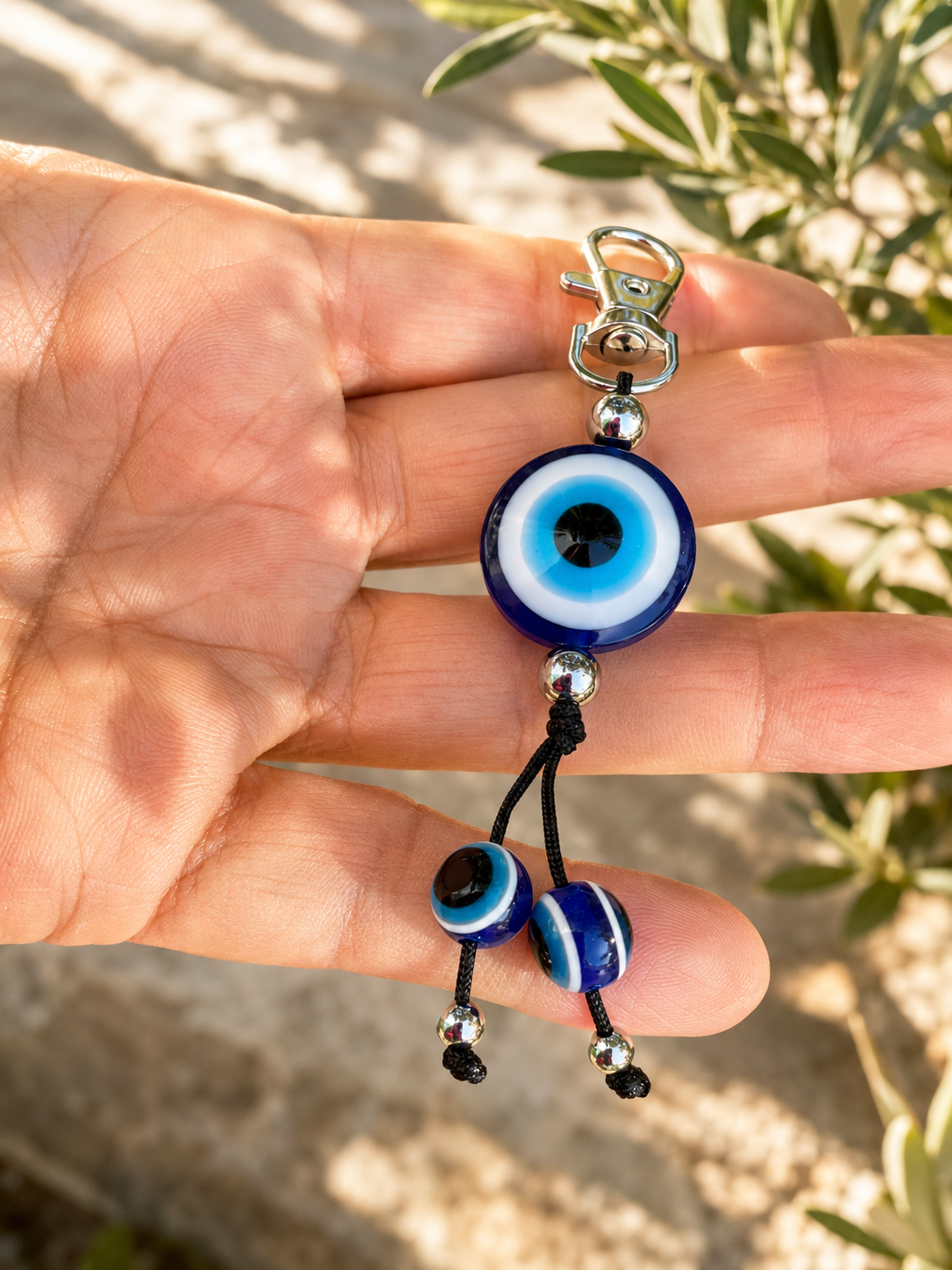 A hand holding a keychain with three blue and white evil eye beads on a black cord, with a silver clasp at the top and a blurred outdoor background.