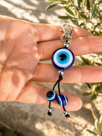 A hand holding a keychain with three blue and white evil eye beads on a black cord, with a silver clasp at the top and a blurred outdoor background.