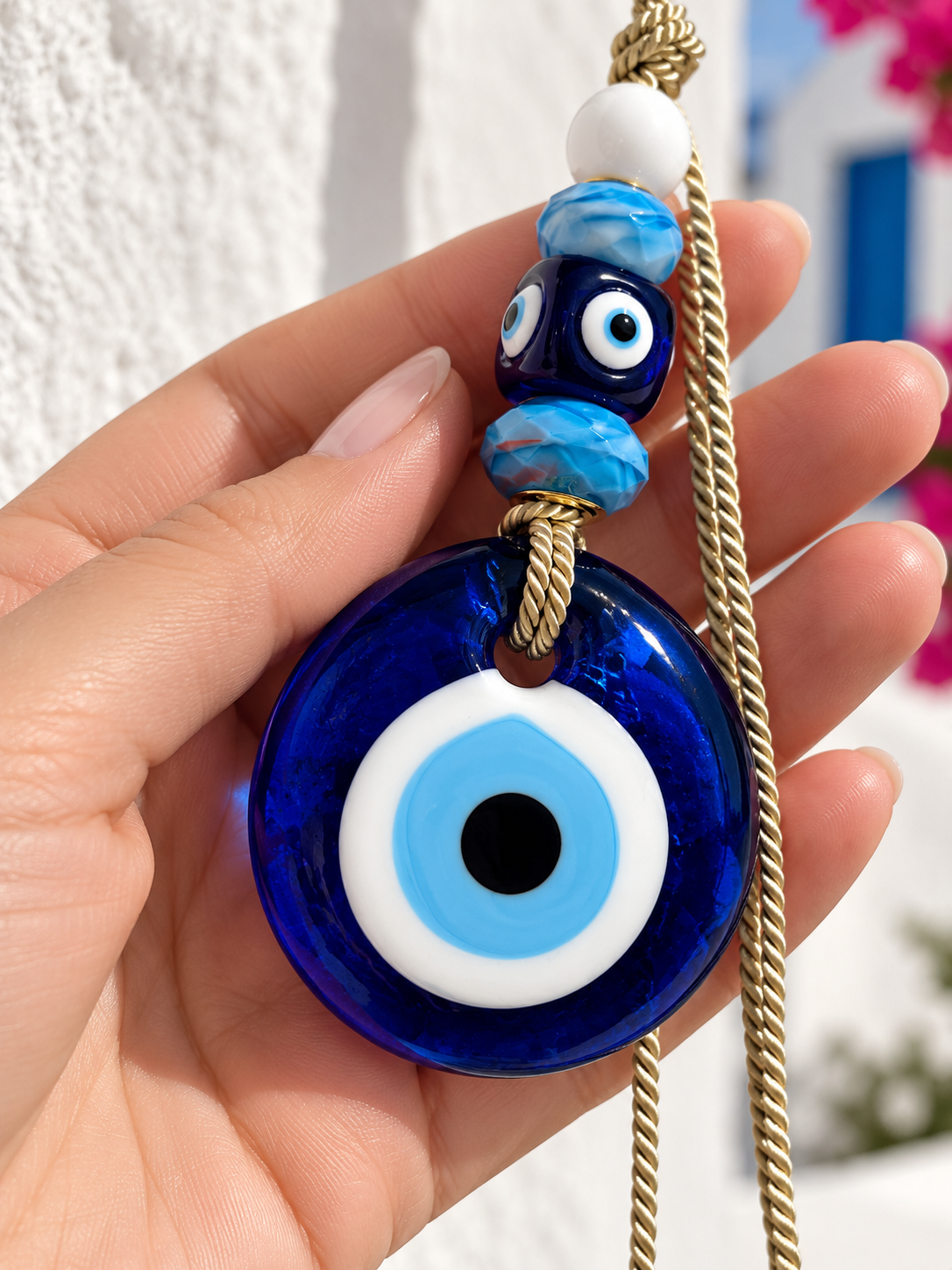 A hand holds a blue evil eye wall hanging charm against a background of white walls and a blue sea.