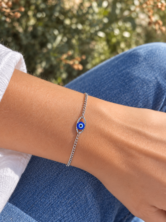 A silver chain bracelet with a blue and white evil eye charm, shown worn on a person's wrist with blue jeans and a white shirt in the background.