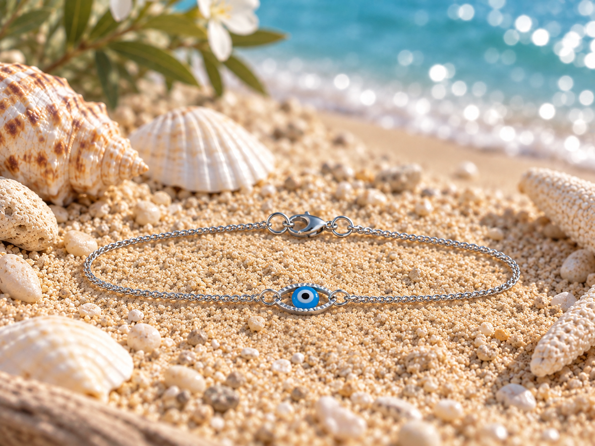 A silver chain bracelet with a blue and white evil eye charm lies on a sandy beach styled with seashells, starfish, and ocean water in the background.