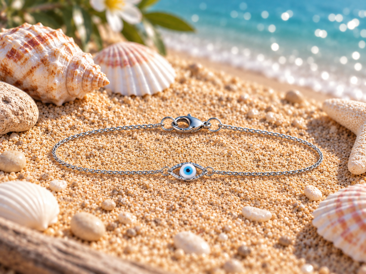 A silver chain bracelet with a blue and white evil eye charm lies on a sandy beach styled with seashells and starfish, with the ocean in the background.